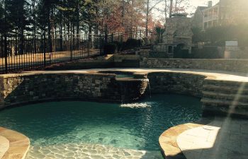 A stone outdoor fireplace by a hardscaped backyard swimming pool with a hot tub and water features installed by Premier Pool Enterprises