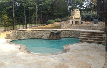 An outdoor stone fireplace built on a deck of a backyard swimming pool with a hot tub and water features installed by Premier Pool Enterprises