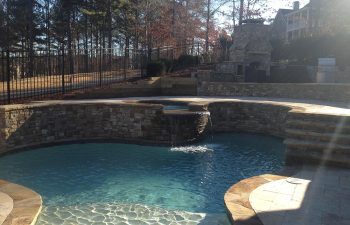 An outdoor stone fireplace built on a deck of a backyard swimming pool with a hot tub and water features installed by Premier Pool Enterprises