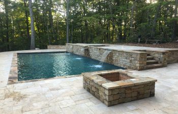 Backyard swimming pool with a water features and a fire pit on a stone deck installed by Premier Pool Enterprises