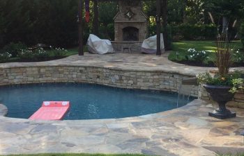 An outdoor stone fireplace built on a deck of a backyard swimming pool with water features installed by Premier Pool Enterprises