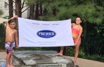 A little boy and a girl in swimming suits standing by a backyard swimming pool and holding a banner with Premier Pool Enterprises logo.