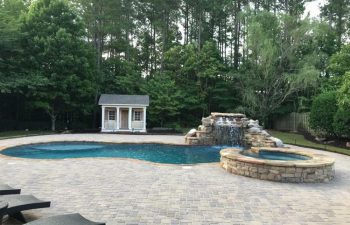Backyard swimming pool with a hardscaped waterfall and a hot tub on a stone deck installed by Premier Pool Enterprises