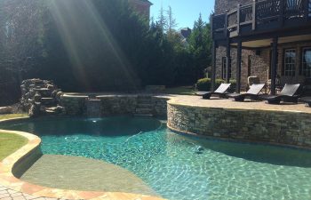 A brick residential house with an outdoor furniture on a stone deck of a swimming pool with a hardscaped waterfall and hot tub installed by Premier Pool Enterprises
