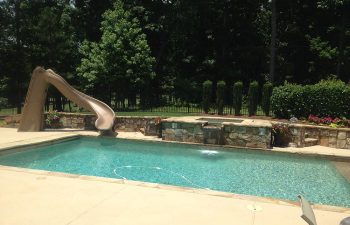 A backyard swimming pool with hardscaped hot tub and a slide installed on the deck.