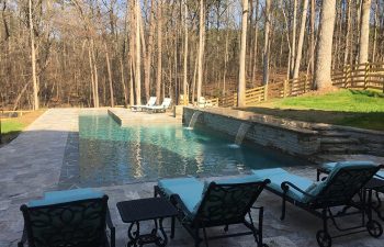 Backyard stone patio with lounge chairs by a swimming pool with water features installed by Premier Pool Enterprises