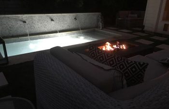 Backyard patio with a fire pit and a swimming pool with water features at night.