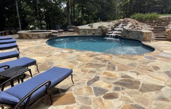 pool furniture on a flagstone deck of a backyard swimming pool with hot tub and hardscaped waterfall
