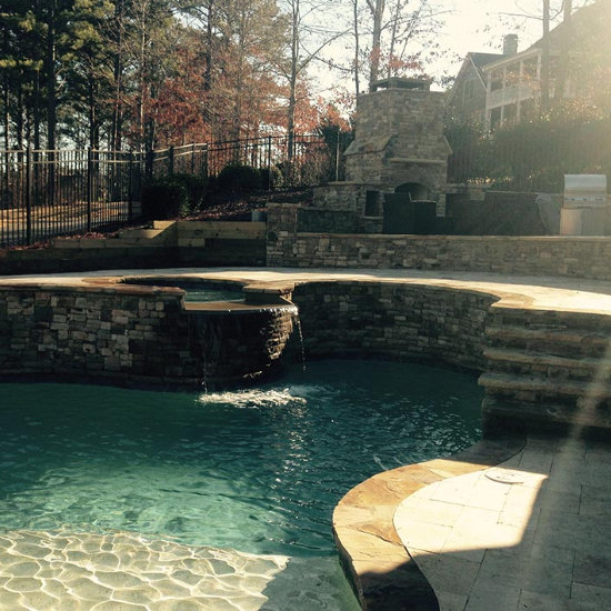 outdoor flagstone patio with a fireplace and and armchairs by a backyard swimming pool with hot tub and waterfall