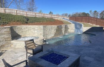 Swimming pool with a hardscaped hot tub and waterfalls installed by Premier Pool Enterprises and Garden chairs around a stone fire pit